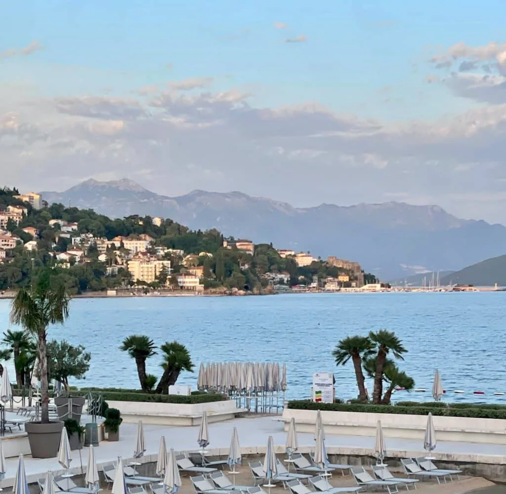 View from a hotel room shows a courtyard with sun loungers, and behind the bay waters. In the distance are mountains.