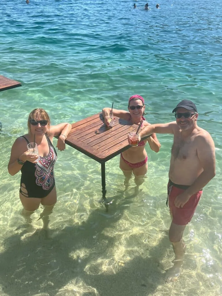 Three tourists standing in water enjoying a cocktail. A table is provided in the water to place drinks upon.