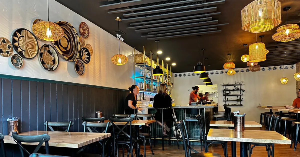 A dining area with woven baskets hanging on a wall and woven lights hanging from the ceiling. Staff at bar with customers.