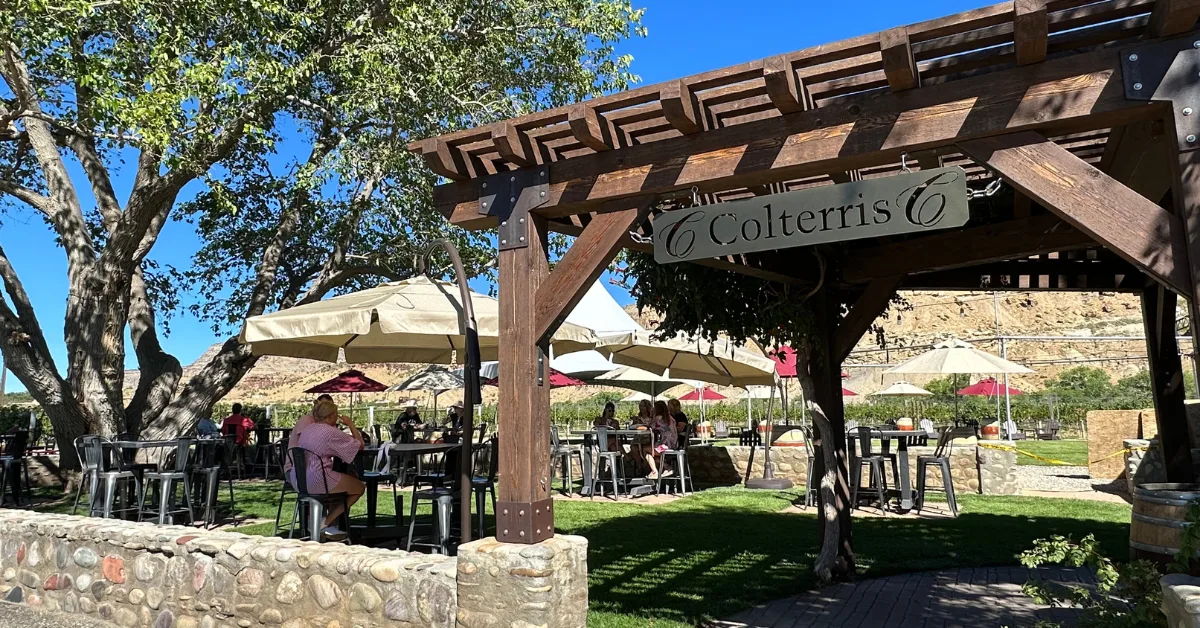 Wooden gazebo entrance to Colterris winery in Palisade and people are sitting wine tasting at outdoor tables with umbrellas as shade.