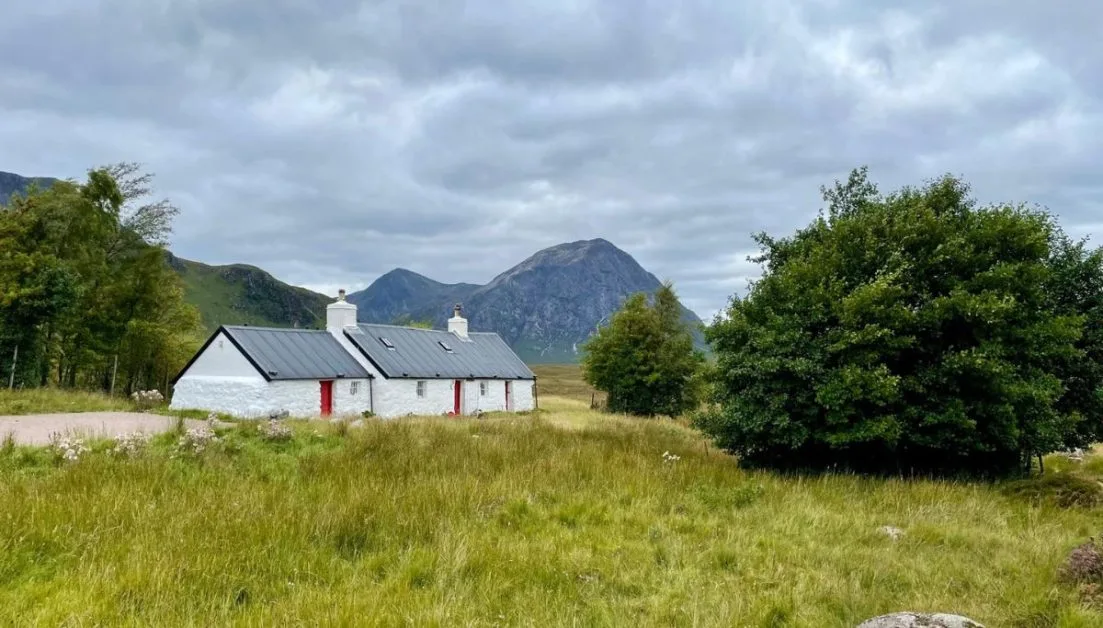 Whitewashed stone cottage in Glencoe Region of Scotland with mountain peaks behind it.