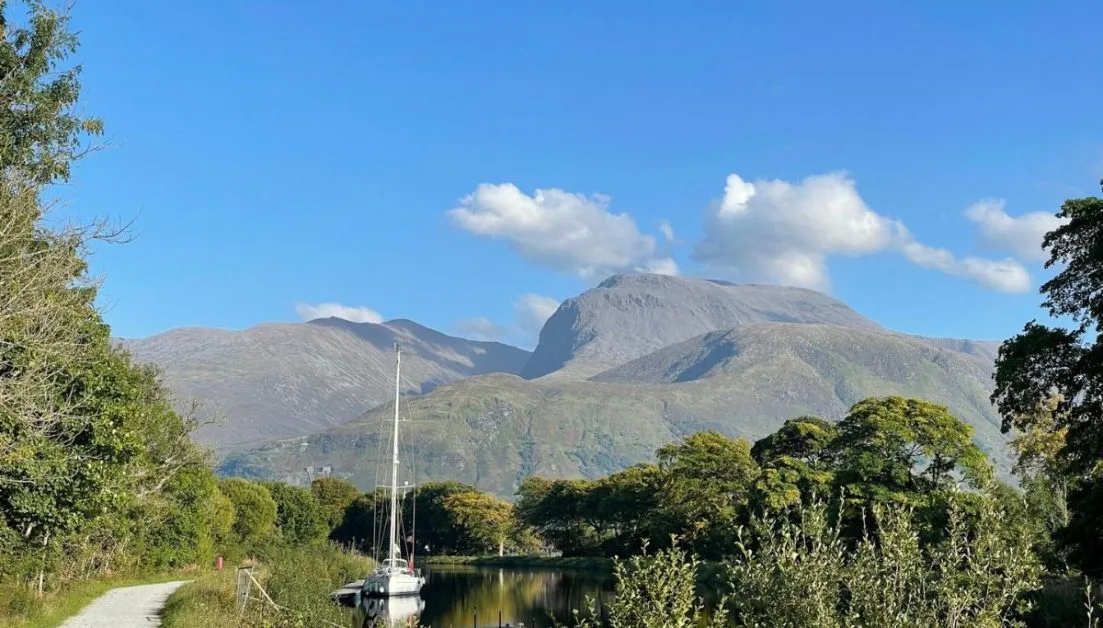 Sail boat is moored alongside a pathway near Fort William. Trees and Glencoe Mountain surround the area.
