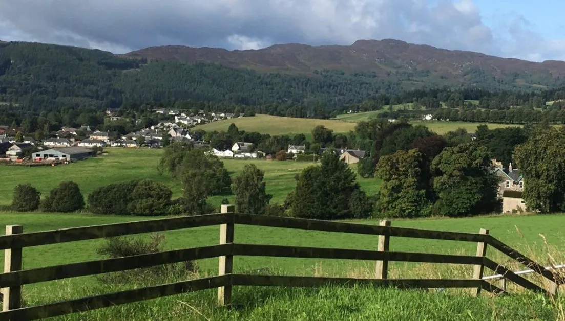 A wooden fence divides farming lands in Perthshire. In the distance is the town of Pitlochry with white washed houses. Mountains rise above the town popular on any Scotland Road Trip Itinerary.