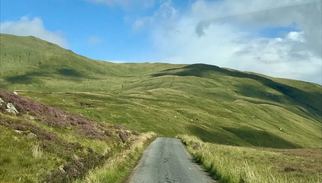 Rural single track road in the western part of scotland surrounded by grassy and heather-clad hills.