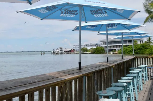 A wooden deck with a line of blue plastic stools that visitors can sit on while eating or drinking. Shaded by blue umbrellas these seats face onto the Sarasota Bay.