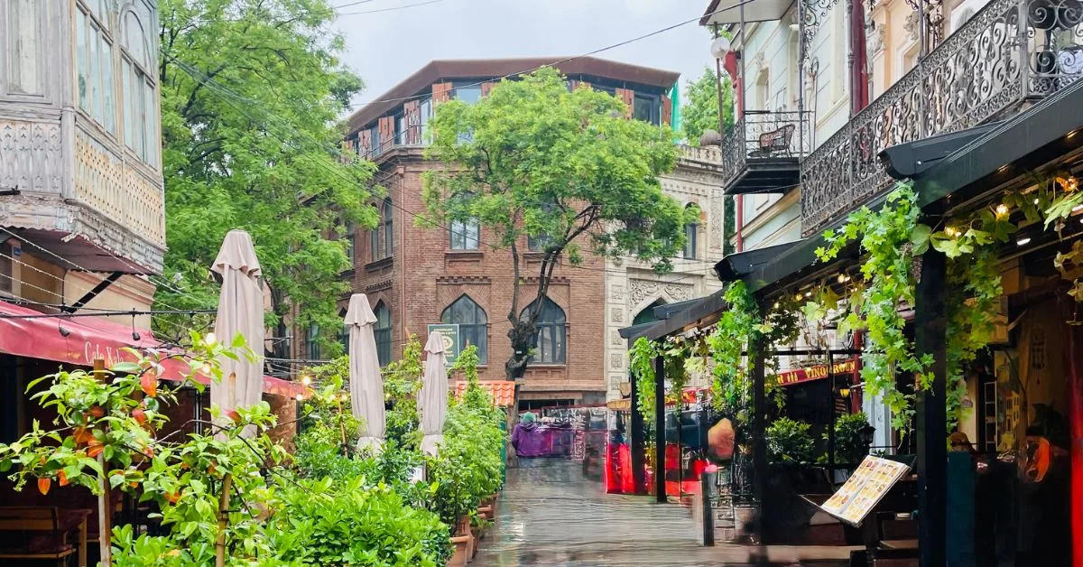 A street lined with cafes and outdoor seating,, shrubs and above is traditional architectural balconies.