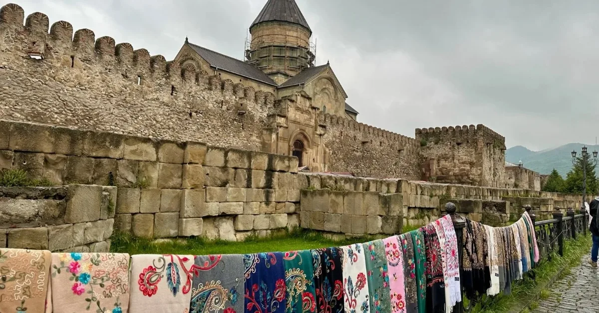 An ancient church in Georgia with an outer wall on which traditional blankets are laid out for sale.