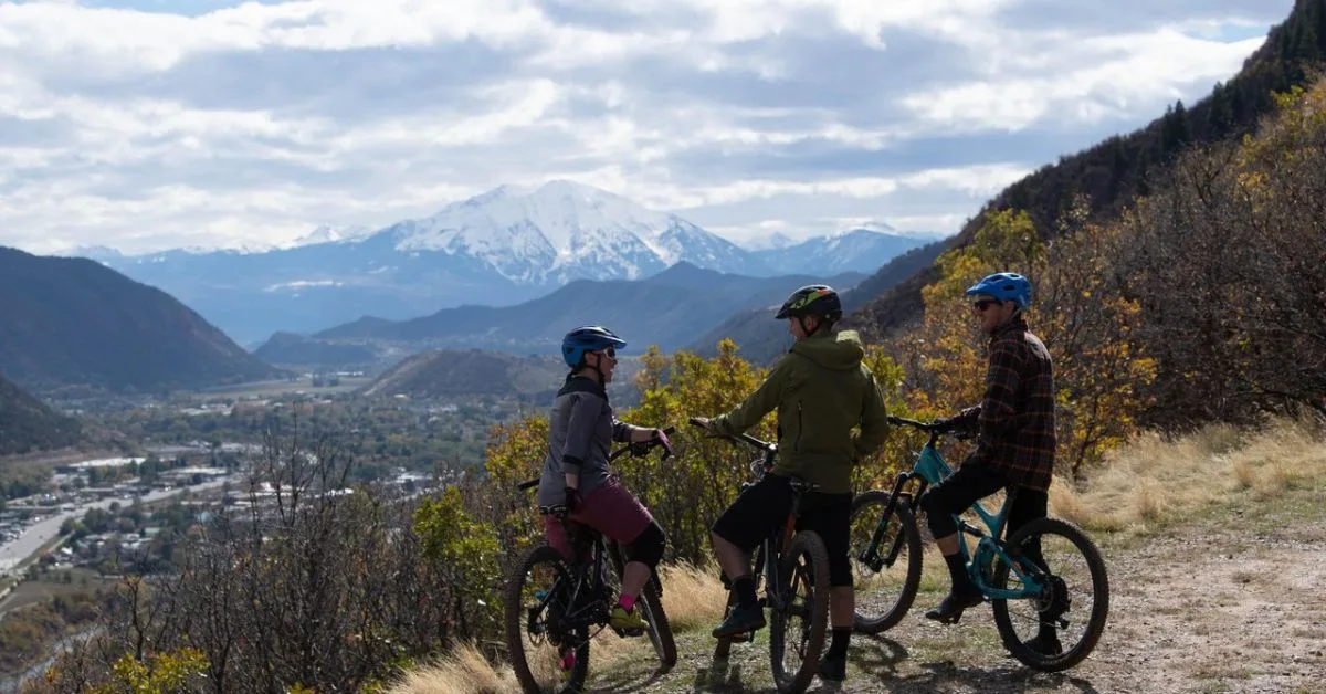 3 cyclists paused looking down at a view of Glenwood Springs town and snow-capped mountains in the distance.
