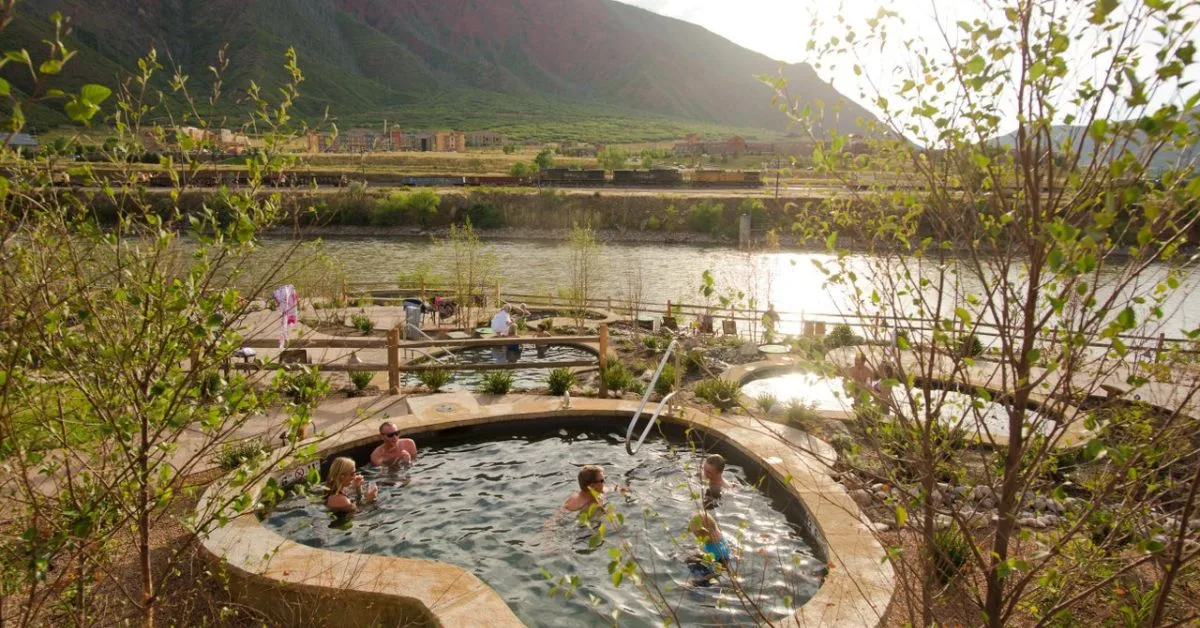 People are soaking in a hot mineral pool surrounded by trees and views of grassy slopes and mountains.