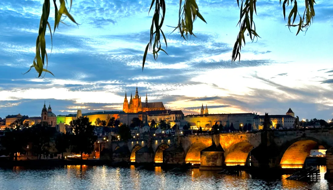 Night time view of Charles Bridge with lights shining on the river below and castle on the hill behind.