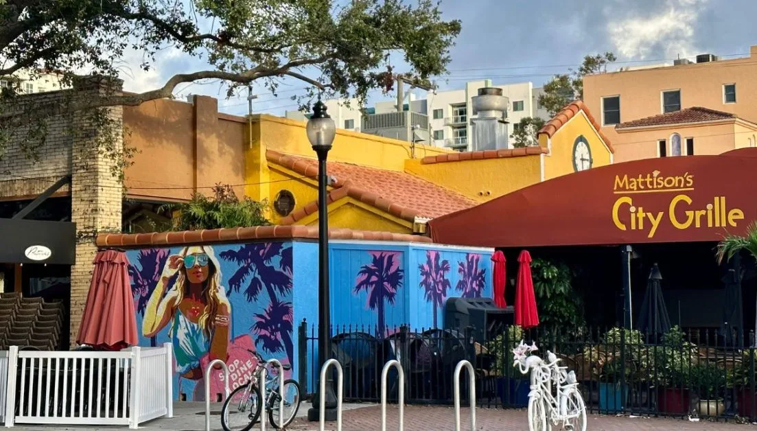 A wall mural with palm trees and a woman shopping brings art to Main Street Sarasota. A restaurant with outdoor seating is adjacent and in front bicycle parking area.