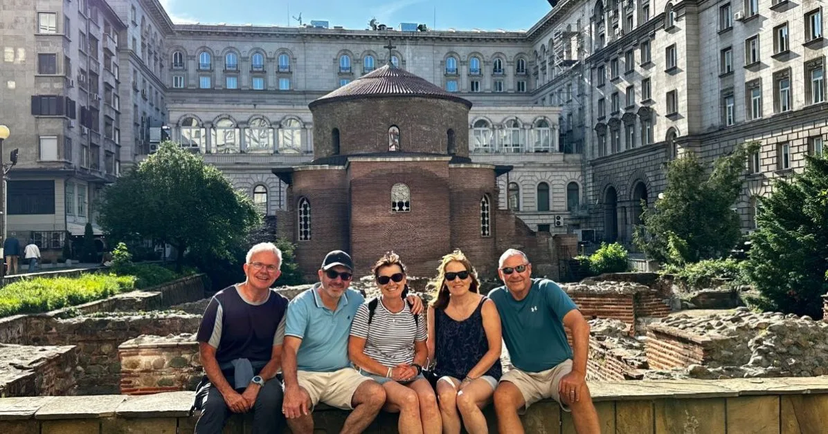 Visitors sitting at the edge of Roman ruins and in front of St George’s red brick Rotunda Church in Sofia. The small church is surrounded grey stone government buildings in contrast.