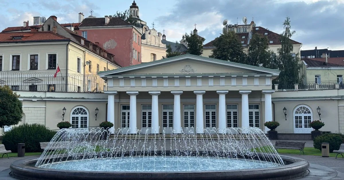 Water fountain sprays into air in front of white pillared palace. Street buildings in the background