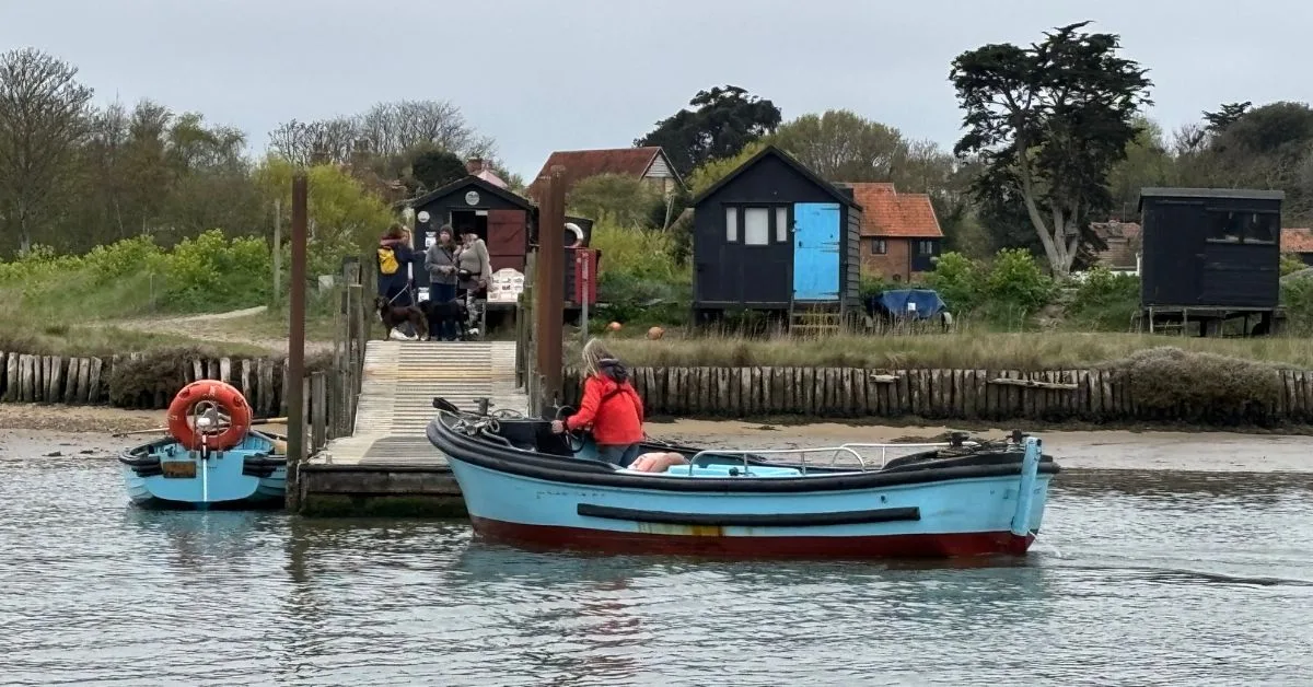 small Ferry boat at boardwalk dock with people waiting to get on the boat. woman in red jacket driving the boat. Wooden huts in the background