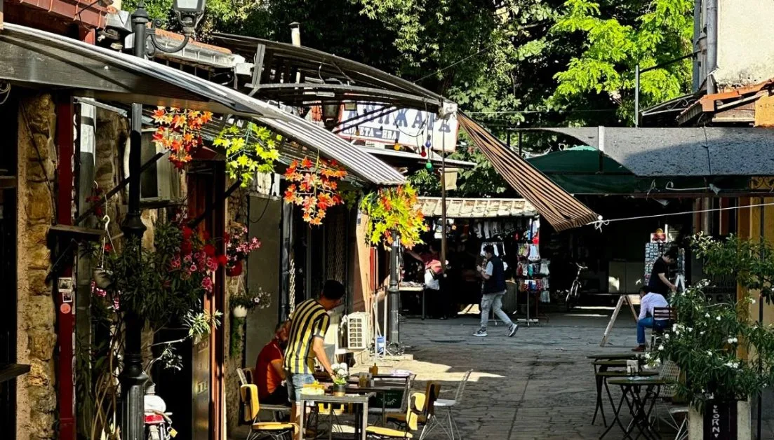 Leafy street with cobbled road. Shops have shaded fronts and it is inviting for people to walk here.