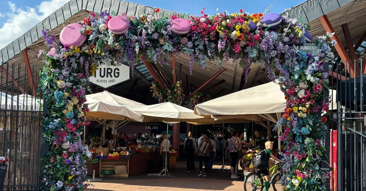 Flower arched entrance to the market with views of some stall inside with canopy parasols for shade as blue skies above