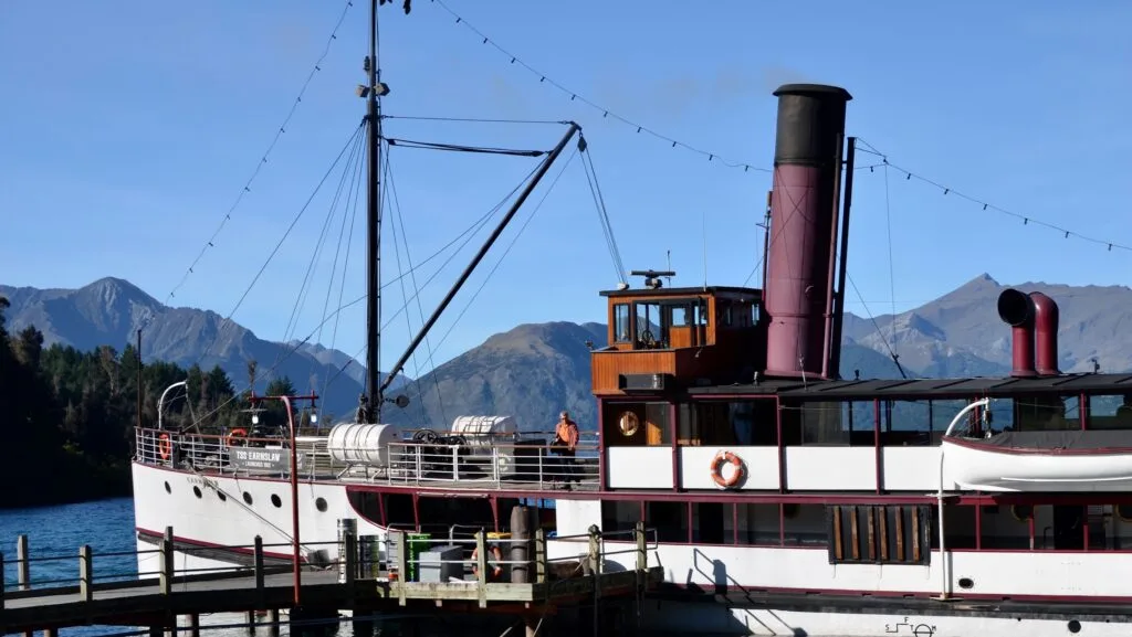 A paddle steamer boat is docked in Queenstown New Zealand. It has a large funnel and rigging and the deck area is open for tourists. The outside of the hull is black and white.