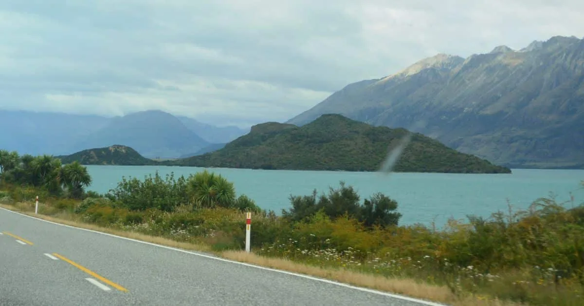 A small road in New Zealand runs next to hills and coastal waterways. The road is small and curves with the coastline.