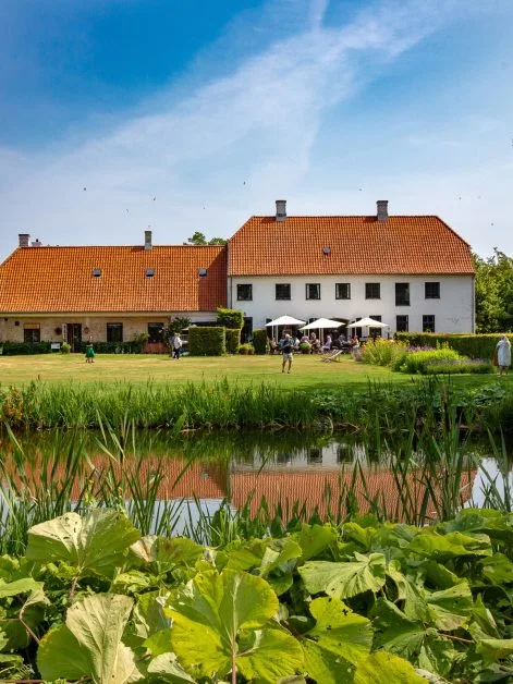 A red tile roof and white painted exterior building with a Lily pond and grass area in front. Several people are sitting under umbrellas at tables.