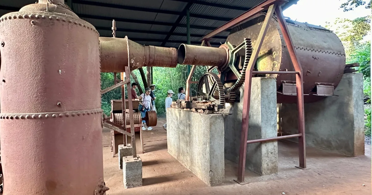 A red metal coffee processing machine with 2 gear wheels. Several tourists are looking at it in Karen Blixen Museum.
