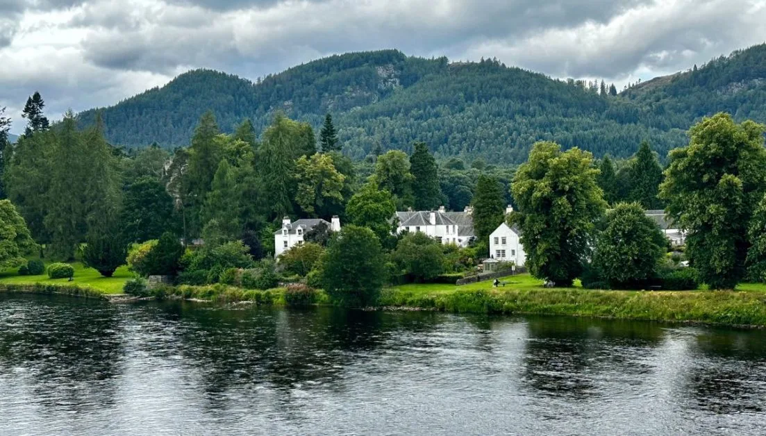 A river with traditional whitewashed stone homes on the banks and thick forest behind it rising up into hill.