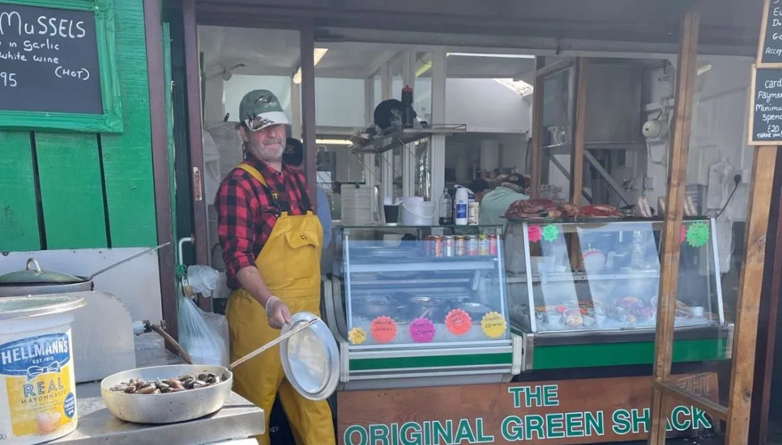 A man dressed in yellow waterproof overalls is cooking fresh seafood at a small outdoor shack in Oban, Scotland. The Original Green Shack.