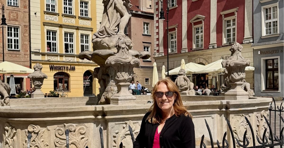 Woman standing in front of the main fountain in the Old Market Square Poznan. Behind her are classic 3-4 story baroque buildings painted in different colours including yellow and red.