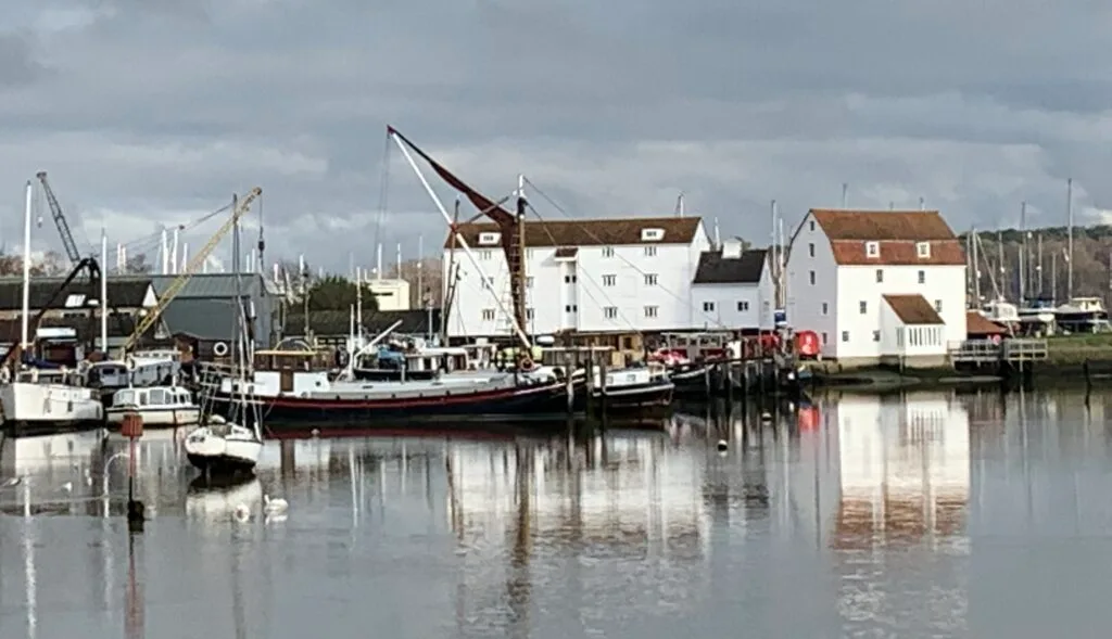 Riverside quay in Woodbridge where house boats and fishing vessels are moored. Old mill with whitewashed cascade sits at the riverside edge. Cranes are located to lift boats in and out of the water.