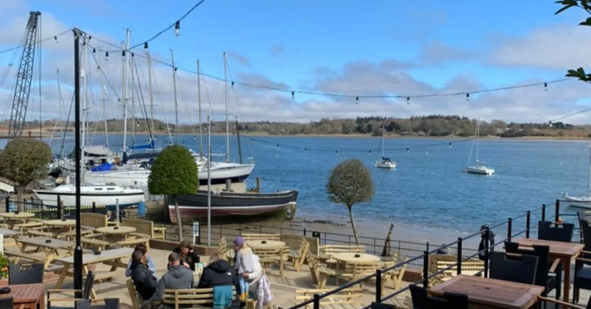Wooden tables and chairs of a pub with views to the river. Sail boats are moored in front of the seating area.