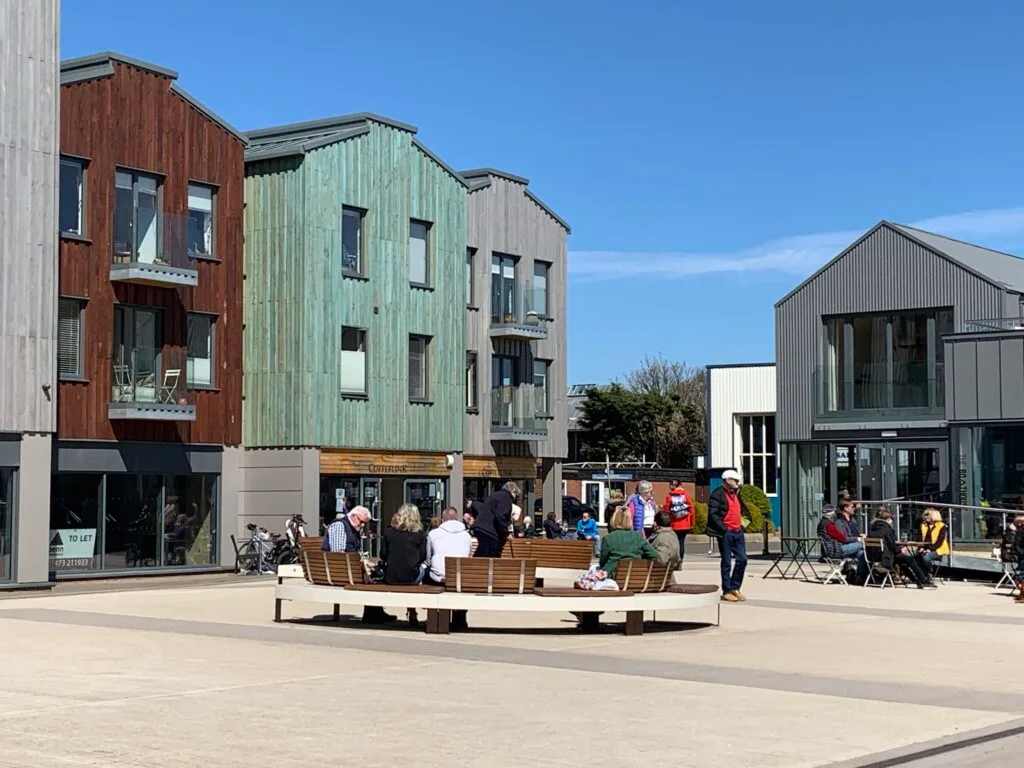 Barn style buildings in brown, green and gray around the Quayside in Woodbridge. On the ground level there are boat yards and people are walking around in the sunshine.