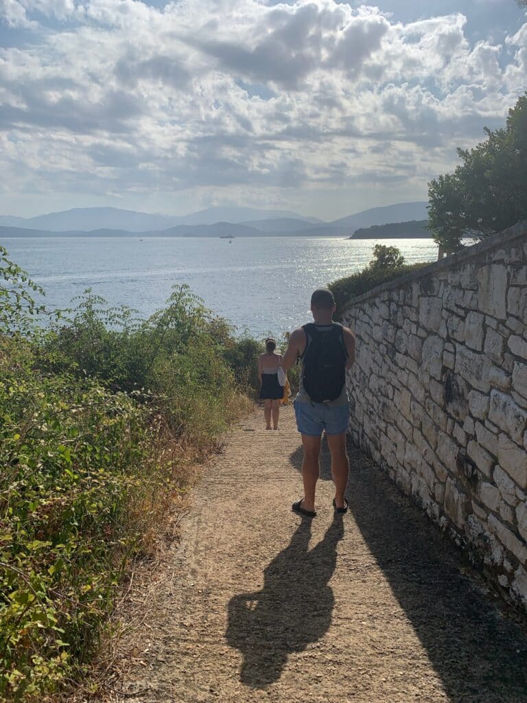 A person is walking down a path with a wall on one side and open scrub land on the other. They are headed to a beach.