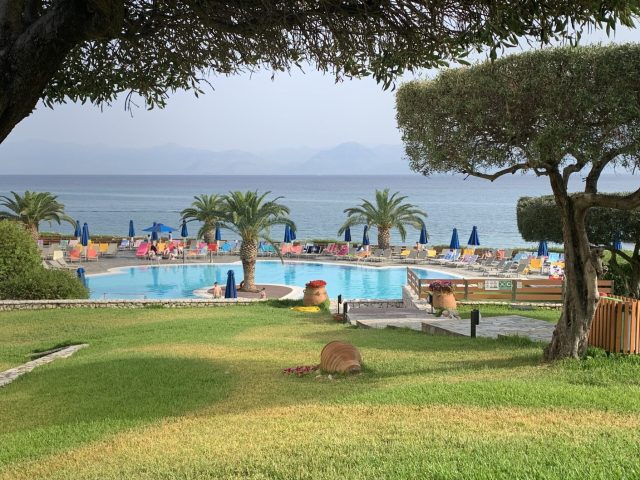 A hotel grounds with grassy areas and a swimming pool. Palm trees and other foliage provides shade.