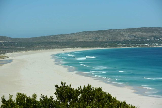 A white sand beach wraps around a bay with trees and bush surrounding it. This beach is in Cape Town area of South Africa.
