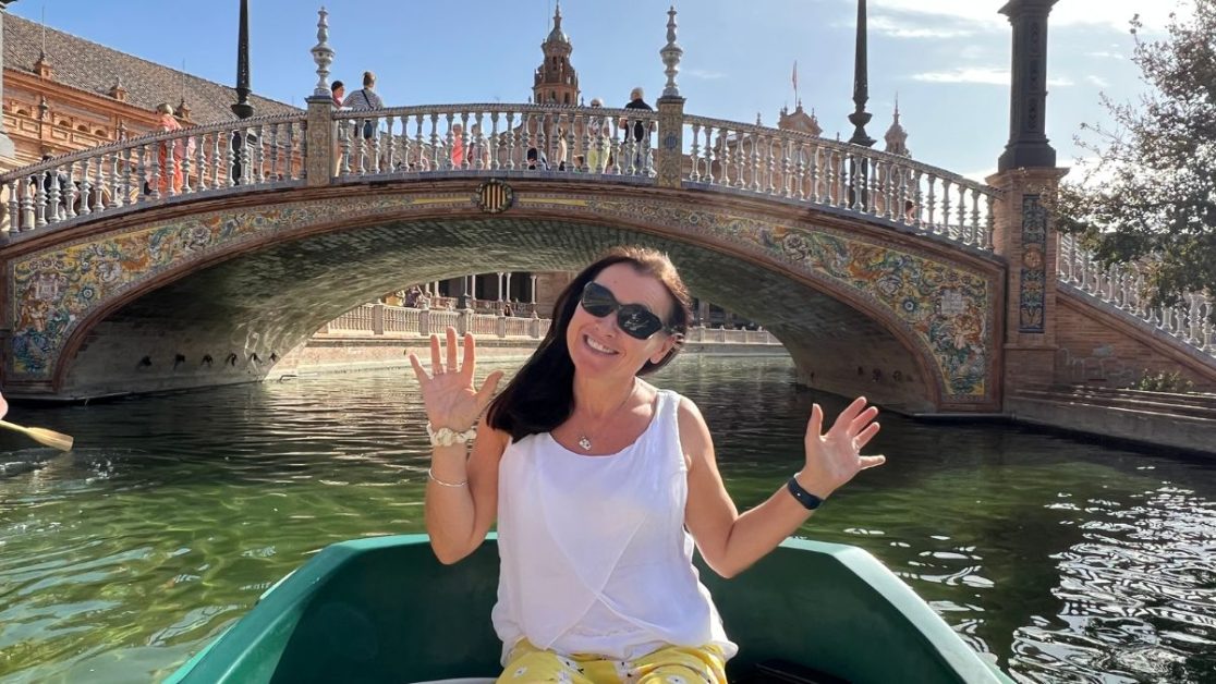 Woman in a small boat exploring canal and bridge system in Seville. The low arched bridge behind her is highly decorated in tiles.