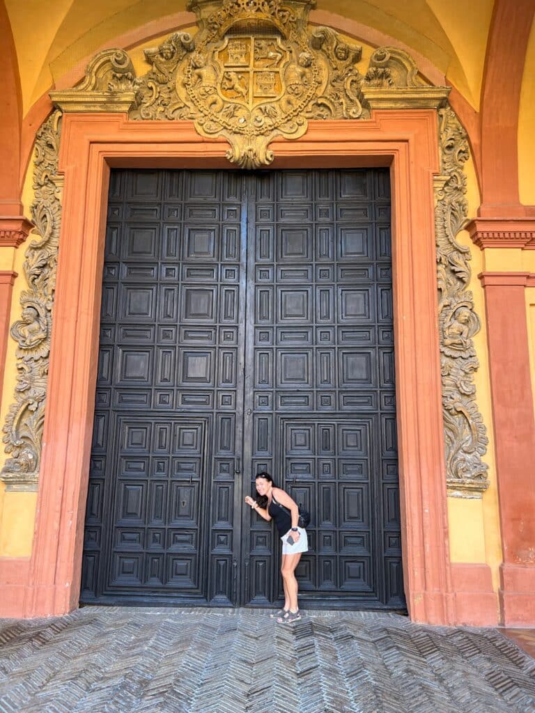 A massive doorway with black doors and an orange decorated wall surrounding it. A woman tourist looks tiny next to the doorway.