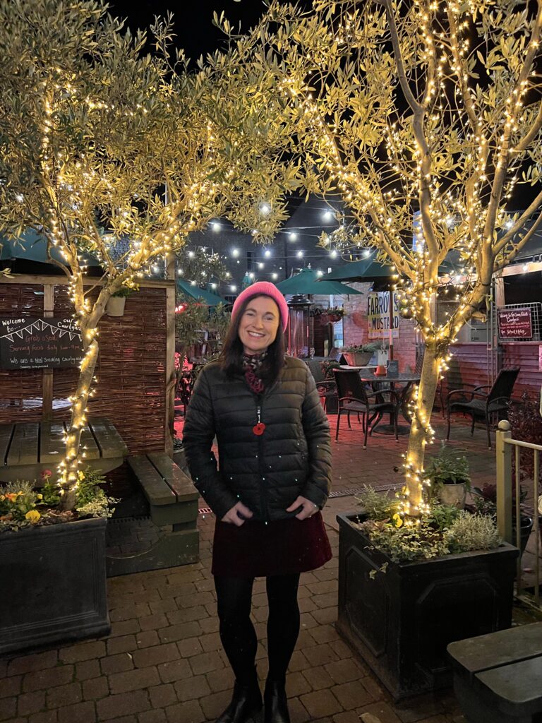 Christmas Lights On Trees shimmer yellow and gold at night. A woman dressed up warm stands in front.