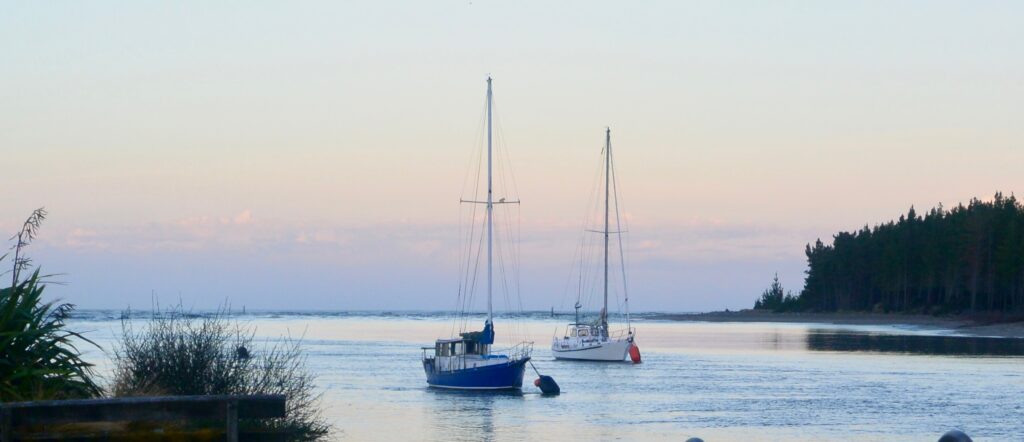 Two sail boats are anchorage in an inlet and beyond are layers of colors of green, pale orange and pink as the sun sets.