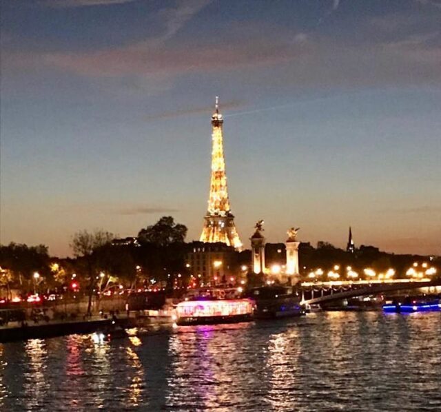 The Eiffel Tower in Paris is lit up and sparkling in the sky as sun is setting. Additional lights from building on each bank illuminate in the water.