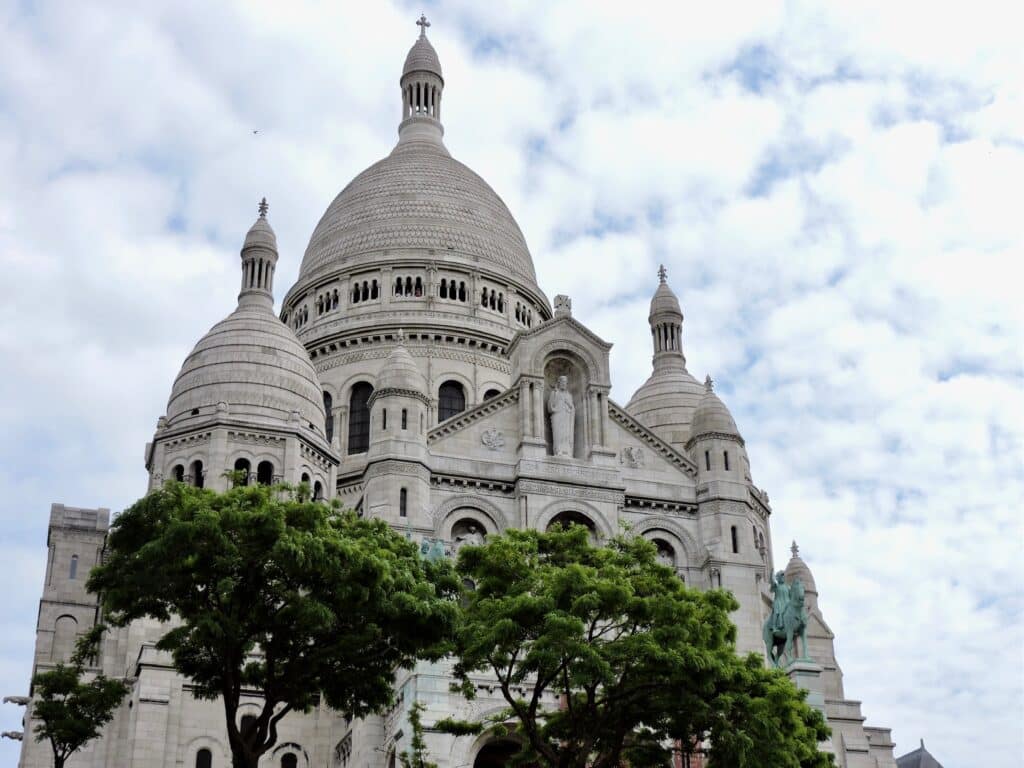 Multiple domes in white create a grand view of the front of Sacre Coeur church in Paris. Trees grow around the terraces in front of the church.