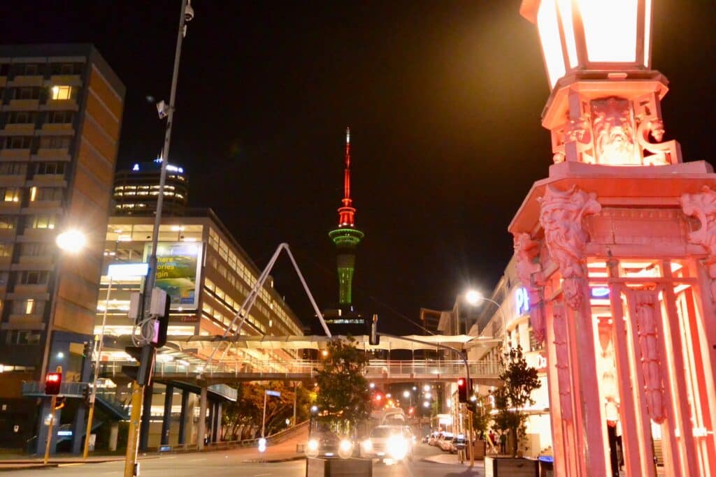 Sky Tower is lit up at night with green lights and red at the tip. Other buildings in central Auckland are also illuminated giving the street a orange/reddish glow.