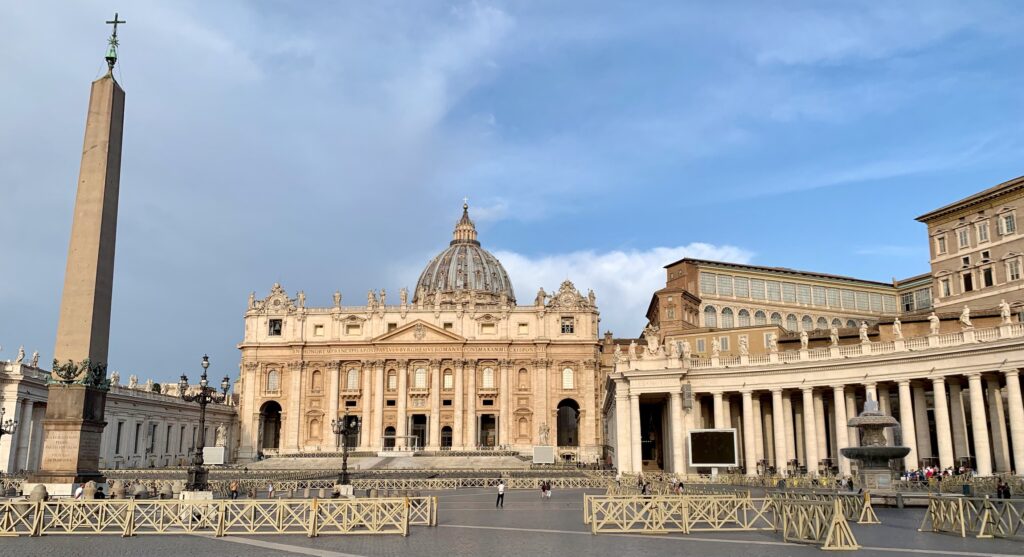 A large square in front of the Vatican City in Rome with an obelisk and curved buildings framed with columns.