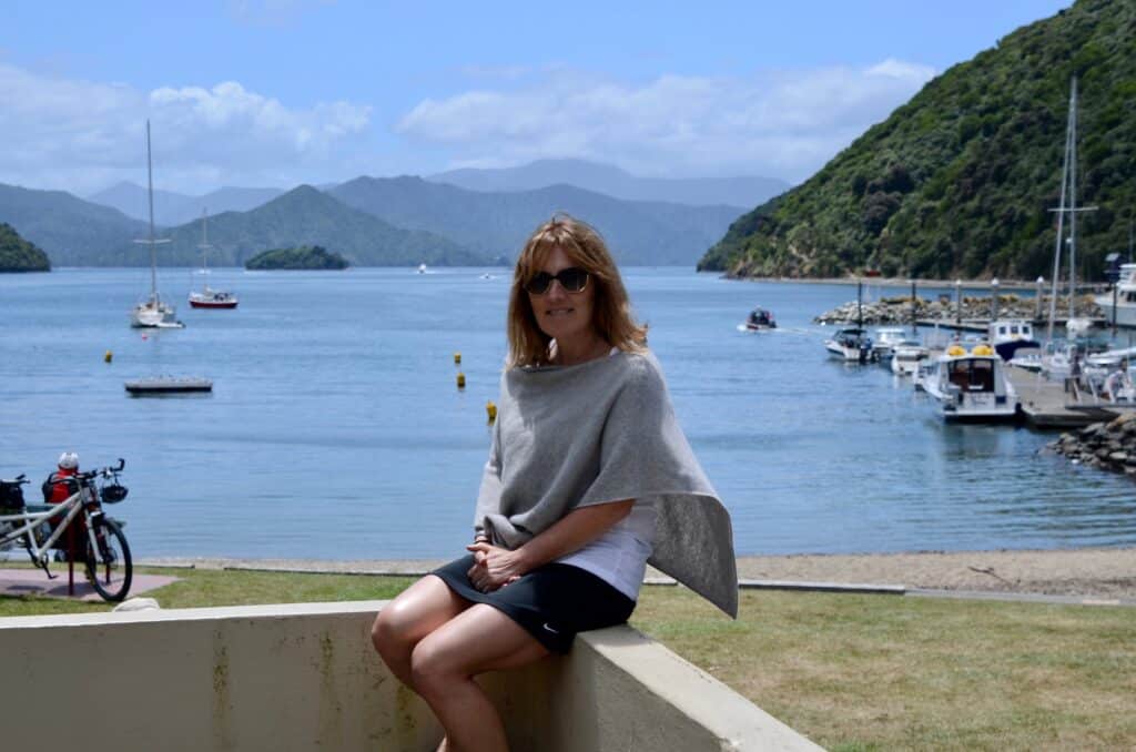 Woman sitting on a wall in a park area in Queen Charlotte. There are fjord type mountains in the background dropping down to the waters. Near to the park a number of sail boats are anchored .