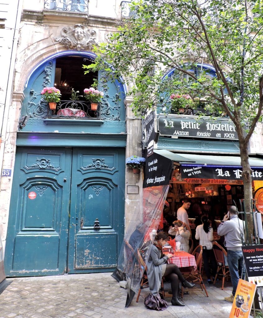a Parisian street with an outdoor cafe next to large blue double doors into a large classic architectural building.
