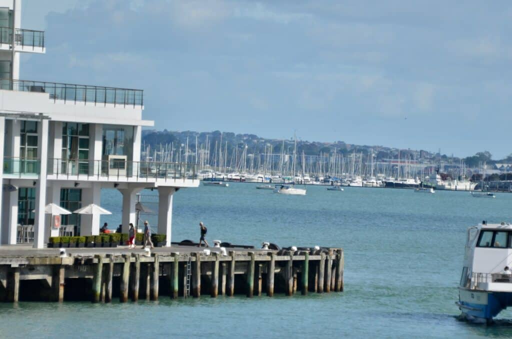 Wooden jetty surrounds a multi-level building with patios on 3 levels. On the bottom level there is a restaurant. The building is next to the calm blue waters of the Marina. In the distance you can see smaller sail boats.