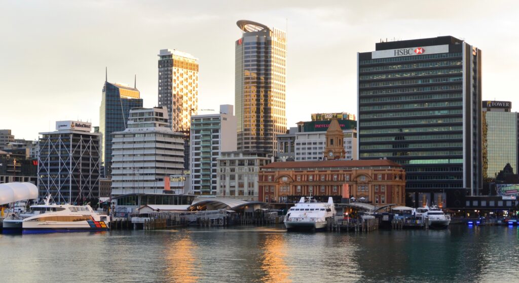 Tall skyscraper buildings and industrial buildings edge the water at a marina/habour area in Auckland. Ferry boats are moored.