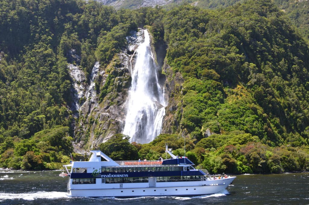 A tour boat sailing in the Milford Sound area of New Zealand and is sailing past a large waterfall.