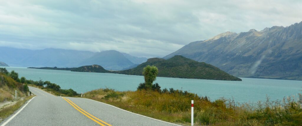 A narrow roadway which hugs a coastline in New Zealand. Mountains can be seen on the far side of the inlet and the water is a light blue colour.