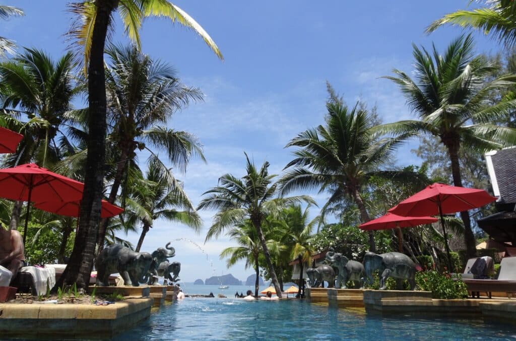 A swimming pool with sun loungers and red parasol shade. Palm trees adorn the area and it looks out to 0pen waters.