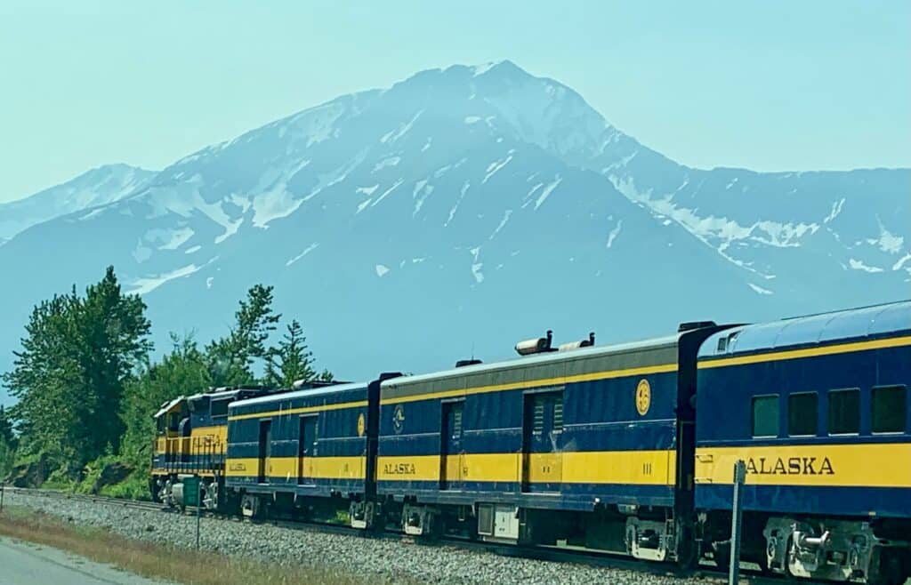 A blue, yellow train with Alaska Railroad signage on a route south with snow peaked mountains in the distance.