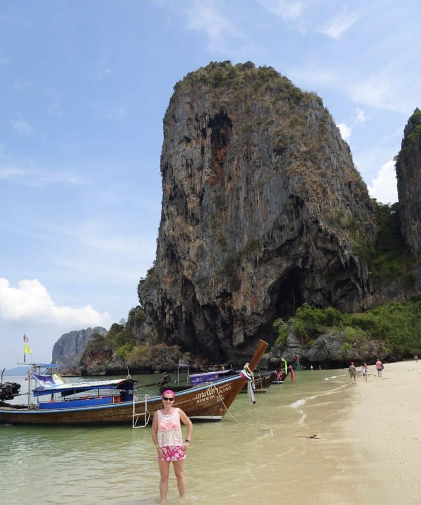 Woman standing on a beach in front of large rock structure. Small boats are anchored on the beach as they transport people from around the islands.